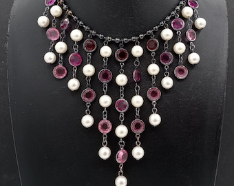 May include: A black and silver necklace with a cascading design of pink and white beads. The necklace features a row of pink beads at the top, followed by a row of white pearls, and then a row of pink beads again. The necklace is finished with a single white pearl at the bottom.