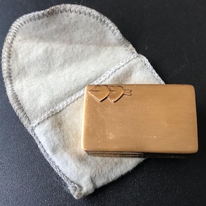 May include: A rectangular, gold-toned metal compact with a heart and arrow design, resting on a light gray pouch. The compact is closed and has a smooth surface. The pouch is made of a soft, felt-like material.