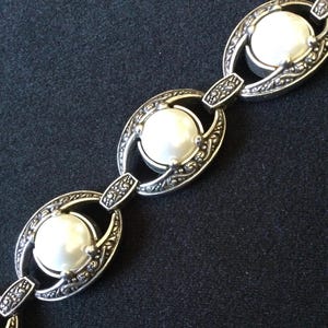 Peut inclure: Un bracelet argent&eacute; compos&eacute; de trois grandes perles rondes blanches, fausses, serties dans des maillons ovales orn&eacute;s. Les maillons sont reli&eacute;s par de plus petits connecteurs d&eacute;coratifs. Le design a une esth&eacute;tique vintage.