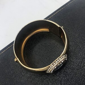 Puede incluir: Un brazalete de oro con un gran grupo de diamantes de imitaci&oacute;n transparentes en un lado. El brazalete est&aacute; abierto y tiene una bisagra.