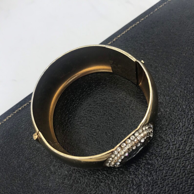 Puede incluir: Un brazalete de oro con un gran grupo de diamantes de imitaci&oacute;n transparentes en un lado. El brazalete est&aacute; abierto y tiene una bisagra.