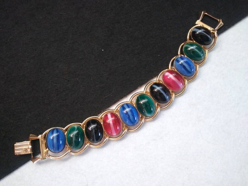 May include: A gold bracelet with oval-shaped stones in shades of blue, green, black, and pink. The stones have a striped pattern.