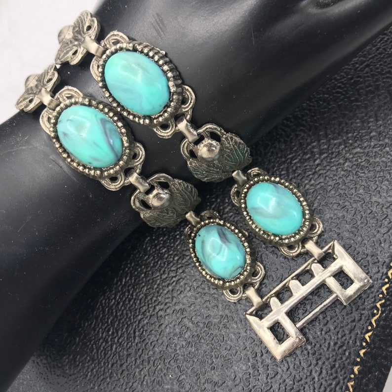 Op de afbeelding: Vintage zilverkleurige armband met ovale turquoise stenen met een gemarmerd effect. De armband heeft sierlijk metaalwerk met blad- en bloemaccenten en een sluiting. Een klassiek sieraad.