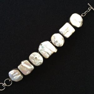 May include: A silver bracelet with irregularly shaped, luminous white pearls. The pearls are connected by silver links and a toggle clasp. The bracelet is displayed against a black background, highlighting the pearls' natural luster.