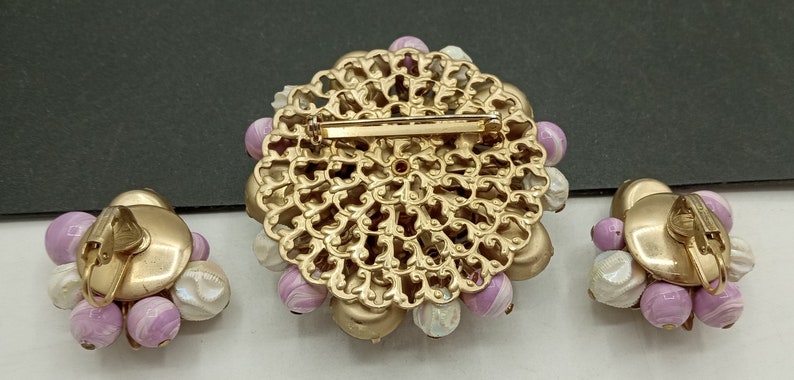 May include: A set of vintage jewelry featuring a large, gold-toned brooch with a filigree design and a cluster of pink and white beads. The set also includes a pair of matching earrings with a similar design.