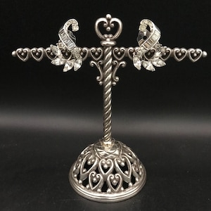 May include: A pair of silver-tone clip-on earrings with clear rhinestones. The earrings are displayed on a silver heart-shaped earring stand.