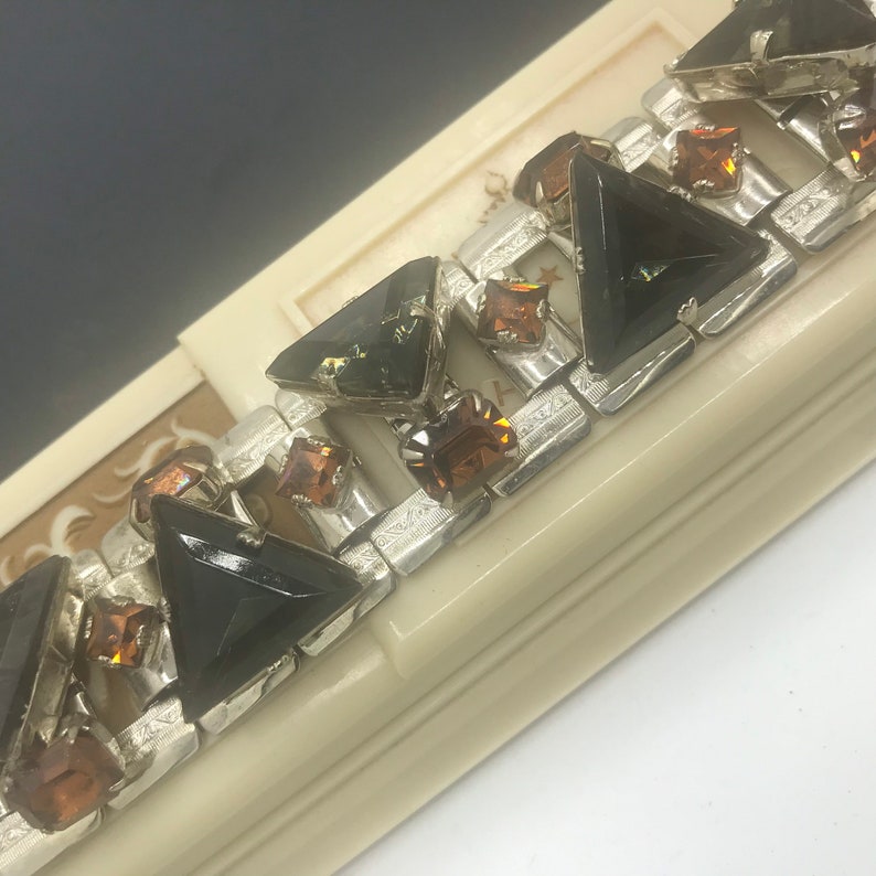 May include: A silver bracelet with black and amber triangular and square shaped rhinestones. The bracelet is in a white box.