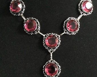 Op de afbeelding: Een zilveren ketting met zeven grote, ronde, roze edelstenen. Elke edelsteen is gezet in een decoratief, geschulpt zilveren frame. De ketting heeft een Y-vormig ontwerp, met een enkele edelsteen hanger in het midden.