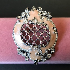 以下が含まれることがあります： ピンクのエナメルデザインとシルバーカラーのメタルフレームが特徴のヴィンテージスタイルのブローチ。ブローチは、ピンクの宝石をあしらった中央の格子模様が特徴で、クリアクリスタルとフローラルアクセントで飾られています。