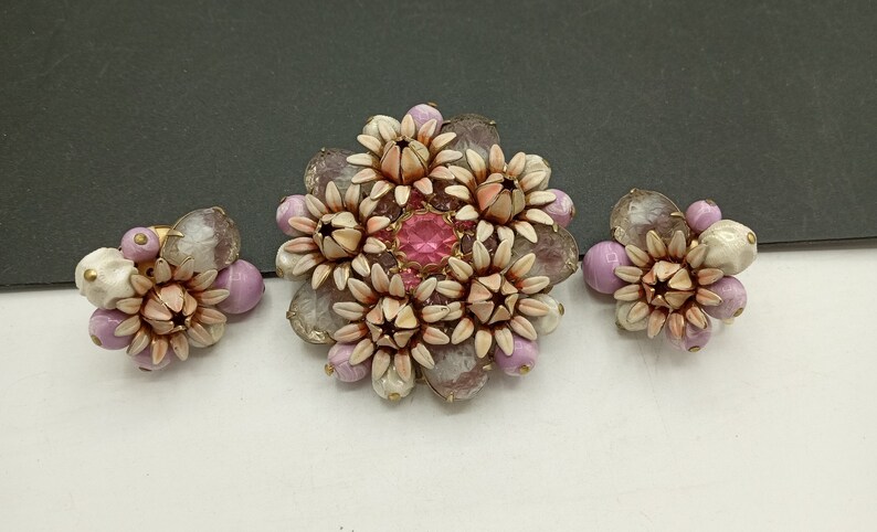 May include: A set of vintage jewelry featuring a large brooch and two earrings. The brooch and earrings are made of white and pink flower-shaped beads with a pink rhinestone in the center of the brooch.
