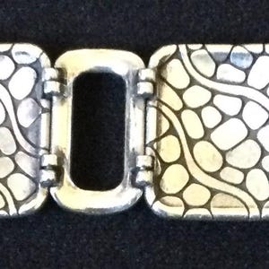 May include: A silver-toned link bracelet featuring four rectangular panels with a raised, textured grid pattern resembling cobblestones or scales, separated by smaller, smooth, open rectangular links and finished with a metal clasp.