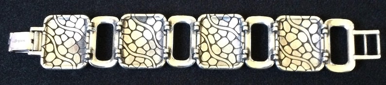 May include: A silver-toned link bracelet featuring four rectangular panels with a raised, textured grid pattern resembling cobblestones or scales, separated by smaller, smooth, open rectangular links and finished with a metal clasp.