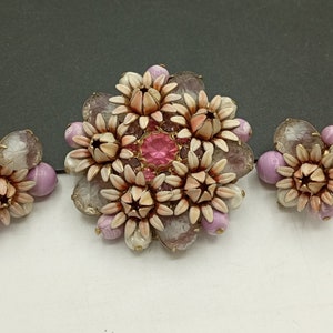 May include: A set of vintage jewelry featuring a large brooch and two earrings. The brooch and earrings are made of white and pink flower-shaped beads with a pink rhinestone in the center of the brooch.