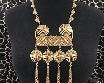 May include: A gold-toned statement necklace with a geometric design. The necklace features a large, rectangular pendant with a textured surface and a series of smaller, circular pendants hanging below.