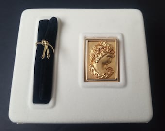 May include: A white velvet jewelry box with a gold-colored metal frame and a raised image of a woman's face. The box has a black velvet lining and a gold-colored metal clasp.