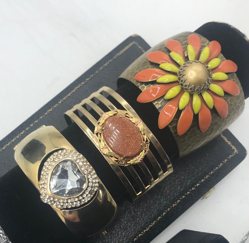 Puede incluir: Tres pulseras doradas con diferentes adornos. La primera pulsera tiene un gran dise&ntilde;o de flor naranja y amarilla. La segunda pulsera tiene una piedra marr&oacute;n engastada en un marco dorado. La tercera pulsera tiene un coraz&oacute;n de cristal transparente.
