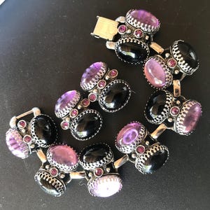 Peut inclure: Un bracelet vintage orn&eacute; de pierres pr&eacute;cieuses ovales noires et violettes serties dans un m&eacute;tal argent&eacute; orn&eacute;. Le bracelet est dot&eacute; d'une fermeture &agrave; fermoir et est pr&eacute;sent&eacute; sur une surface sombre. Les pierres sont rehauss&eacute;es de petites pierres roses rondes.