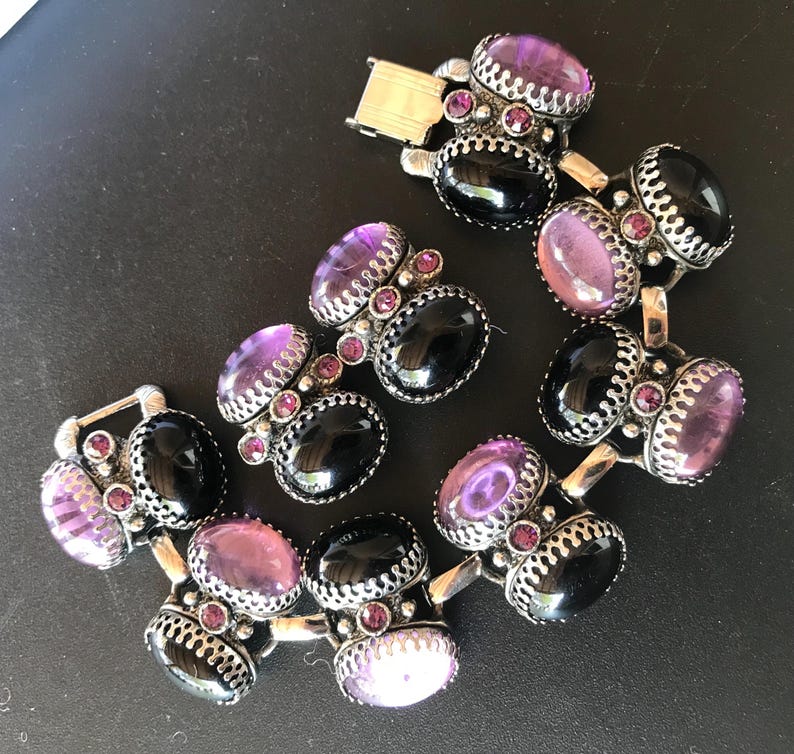 Peut inclure: Un bracelet vintage orn&eacute; de pierres pr&eacute;cieuses ovales noires et violettes serties dans un m&eacute;tal argent&eacute; orn&eacute;. Le bracelet est dot&eacute; d'une fermeture &agrave; fermoir et est pr&eacute;sent&eacute; sur une surface sombre. Les pierres sont rehauss&eacute;es de petites pierres roses rondes.