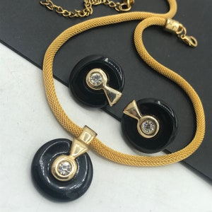 May include: A gold chain necklace with a black pendant featuring a rhinestone. The necklace is paired with matching earrings with black circular pendants and rhinestones.
