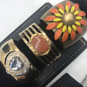 Puede incluir: Tres pulseras doradas con diferentes dise&ntilde;os. Una pulsera presenta una gran flor naranja y amarilla con un centro dorado. Otra pulsera tiene una piedra marr&oacute;n engastada en un marco dorado. La tercera pulsera tiene un coraz&oacute;n de cristal transparente rodeado de cristales m&aacute;s peque&ntilde;os.