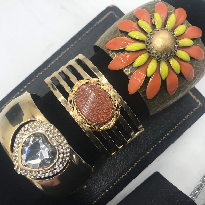 Puede incluir: Tres pulseras doradas con diferentes dise&ntilde;os. Una pulsera presenta una gran flor naranja y amarilla con un centro dorado. Otra pulsera tiene una piedra marr&oacute;n engastada en un marco dorado. La tercera pulsera tiene un coraz&oacute;n de cristal transparente rodeado de cristales m&aacute;s peque&ntilde;os.