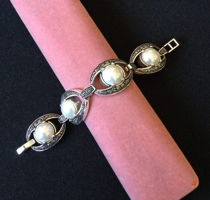 Peut inclure: Un bracelet argent&eacute; orn&eacute; de grandes perles blanches rondes, serties dans des maillons ovales d&eacute;coratifs. Le bracelet pr&eacute;sente un motif orn&eacute; et une fermeture &agrave; fermoir. Le bijou est pr&eacute;sent&eacute; sur une surface rose cylindrique sur fond noir.