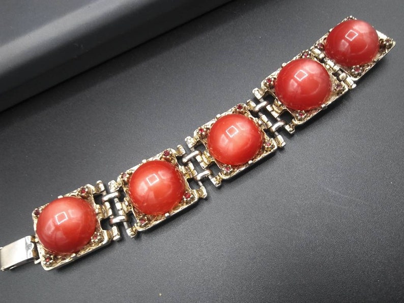 May include: A vintage bracelet featuring five large, round, coral-colored cabochons set in gold-toned metal squares. The squares are connected by a silver-toned chain. Small red rhinestones accent the edges of the cabochon settings.