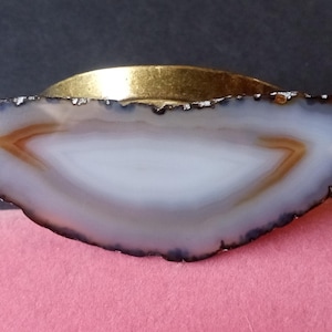 May include: A white agate slice with brown banding, set in a gold-toned metal frame. The agate is a natural stone with a unique pattern.
