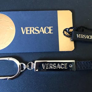 May include: A black leather keychain with a silver-tone metal plate engraved with the word "VERSACE". A matching black tag with "VERSACE" is attached. A blue and gold card with the brand name is also present.