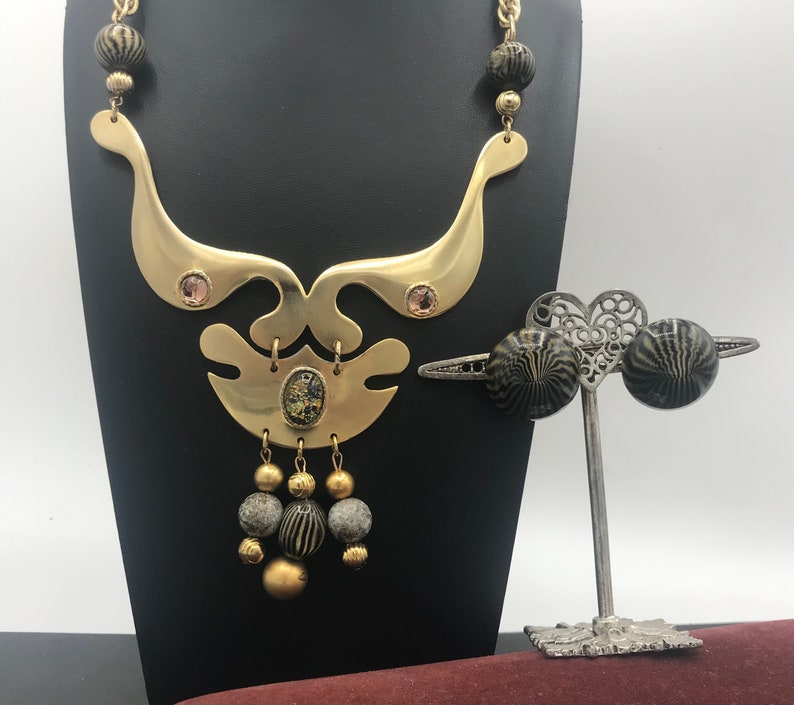 May include: A gold necklace with a geometric design and black and gold beads. The necklace is on a black stand. A pair of black and gold earrings are on a silver stand with a heart-shaped design.