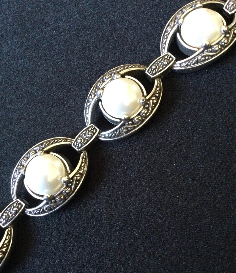 Peut inclure: Un bracelet argent&eacute; compos&eacute; de trois grandes perles rondes blanches, fausses, serties dans des maillons ovales orn&eacute;s. Les maillons sont reli&eacute;s par de plus petits connecteurs d&eacute;coratifs. Le design a une esth&eacute;tique vintage.