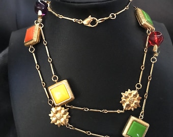 Puede incluir: Collar dorado con detalles cuadrados y redondos de colores. El collar presenta piedras naranjas, amarillas, verdes, rojas y moradas, junto con detalles florales y de cadena dorados. El collar se exhibe en un maniquí negro.