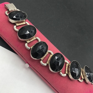 May include: A silver-toned bracelet with large, faceted black stones. The oval-shaped stones are set in silver and connected by silver links. The bracelet is displayed on a pink surface.