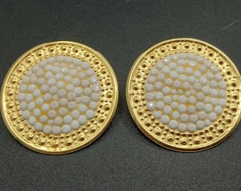 Op de afbeelding: Een paar gouden oorbellen met een wit, rond ontwerp. De oorbellen zijn rond en hebben een getextureerde rand.