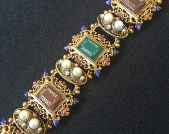 Puede incluir: Pulsera dorada con eslabones rectangulares y ovalados alternados. Los eslabones rectangulares muestran piedras marrones y verdes, mientras que los eslabones ovalados presentan perlas de imitación. La pulsera está adornada con detalles rosas, azules y blancos, creando una estética vintage.