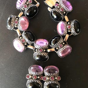 Peut inclure: Un ensemble de bijoux vintage comprenant un bracelet et des boucles d'oreilles assorties. Le bracelet et les boucles d'oreilles ont des pierres ovales noires et violettes, rehauss&eacute;es de petites pierres roses et d'&eacute;l&eacute;ments m&eacute;talliques argent&eacute;s.