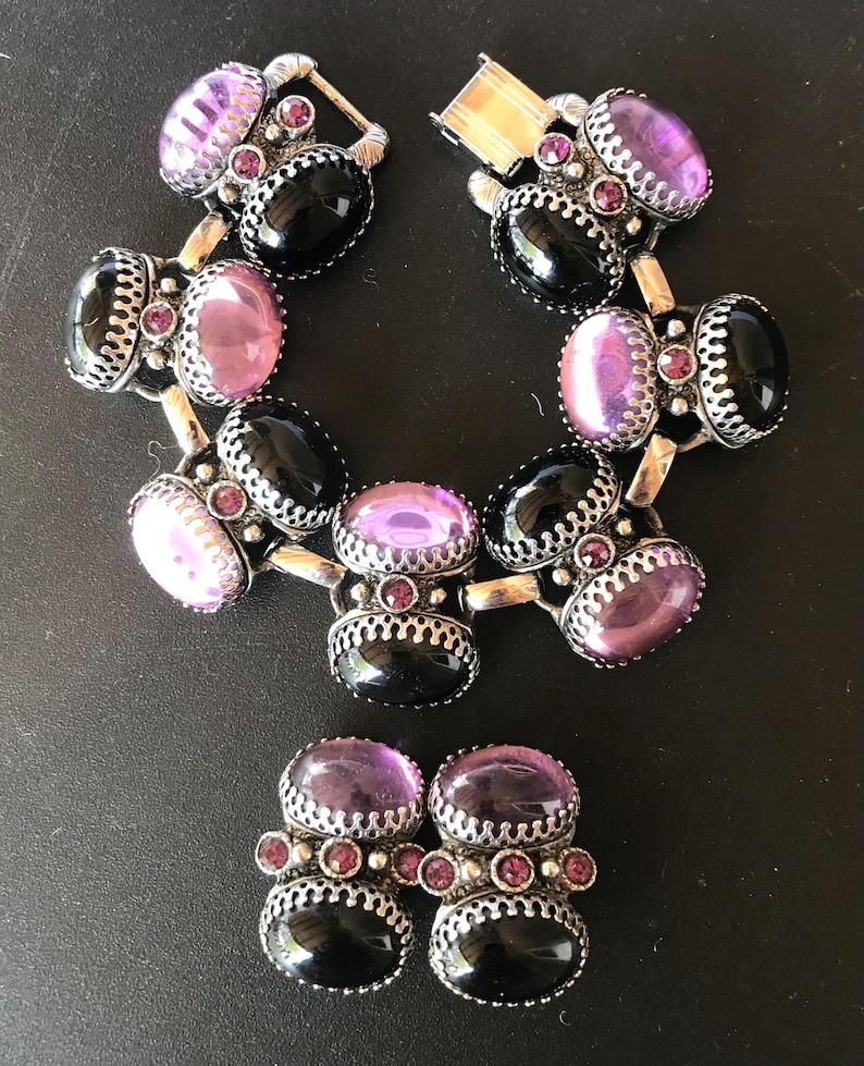Peut inclure: Un ensemble de bijoux vintage comprenant un bracelet et des boucles d'oreilles assorties. Le bracelet et les boucles d'oreilles ont des pierres ovales noires et violettes, rehauss&eacute;es de petites pierres roses et d'&eacute;l&eacute;ments m&eacute;talliques argent&eacute;s.