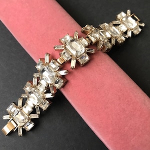 May include: A gold bracelet with a geometric design featuring clear, faceted crystals. The bracelet is made of metal and has a clasp closure.