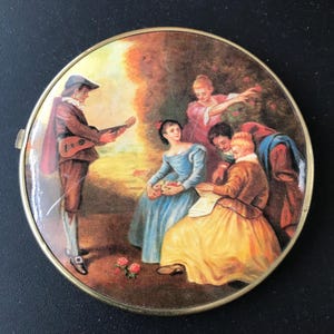 May include: A round, vintage compact with a painted scene of a man playing a lute for a group of people in a garden. The compact has a gold-colored metal frame and a clasp. The scene features figures in period clothing, with a warm color palette.