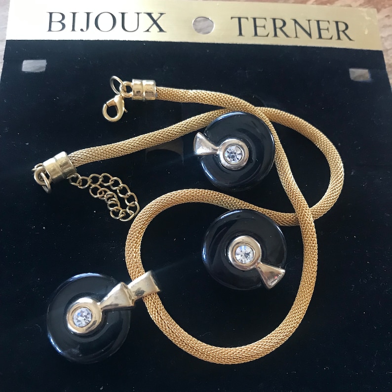 May include: A gold chain necklace and earrings set with black and gold round pendants featuring a clear rhinestone in the center. The jewelry is displayed on a black background with the text "Bijoux Terner".