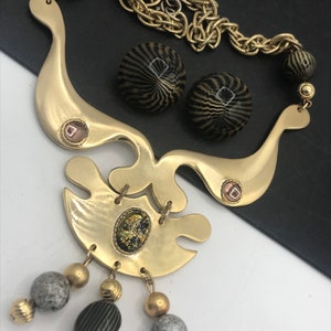 May include: A gold tone necklace with a geometric design and black and gold patterned beads. The necklace has a pendant with a gold tone design and a small, clear stone.
