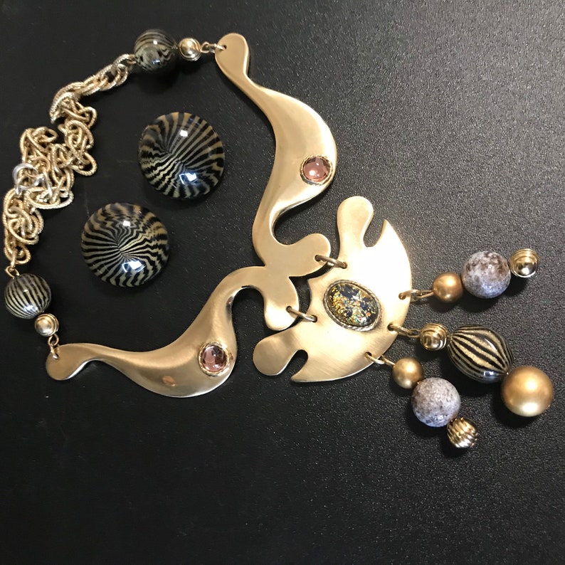 May include: A gold necklace with a large abstract pendant and a chain. The pendant has a black and gold design and is accented with small, round, clear stones. The necklace also has a small, round, black and gold earring.