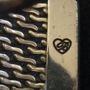 May include: Close-up of a gold and silver-toned metal bracelet clasp. The clasp has a rectangular shape with a heart-shaped logo. The bracelet features a chain-link design.