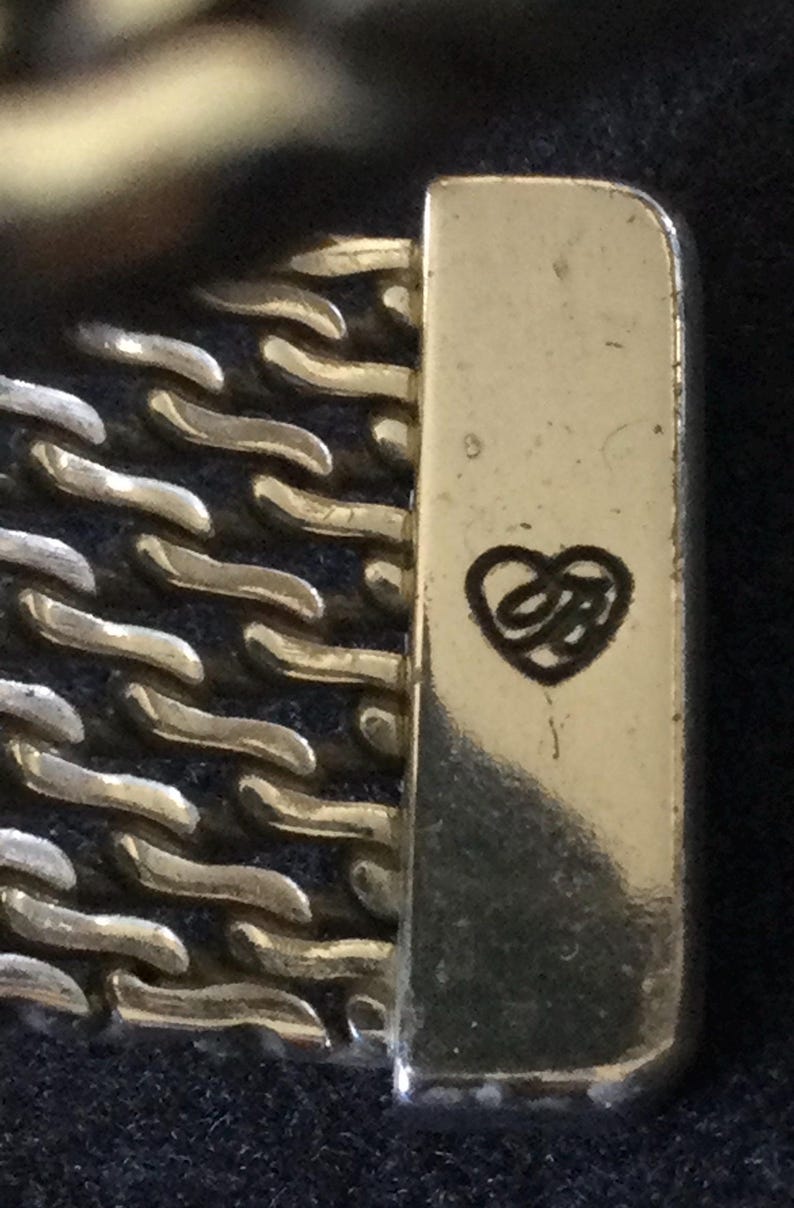 May include: Close-up of a gold and silver-toned metal bracelet clasp. The clasp has a rectangular shape with a heart-shaped logo. The bracelet features a chain-link design.