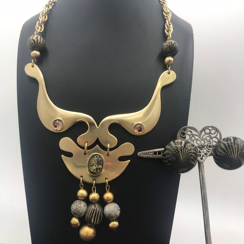 May include: A gold necklace with a geometric design and black and gold beads. The necklace has a pendant with a large, round, gold-colored stone. The necklace is on a black background.