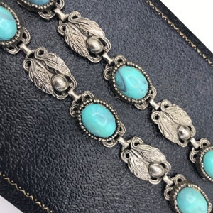 Op de afbeelding: Close-up van een vintage zilverkleurige armband met ovale turquoise stenen. De armband heeft sierlijke blad- en kraaldetails. Het ontwerp is klassiek en elegant. De armband wordt gepresenteerd op een zwarte ondergrond.
