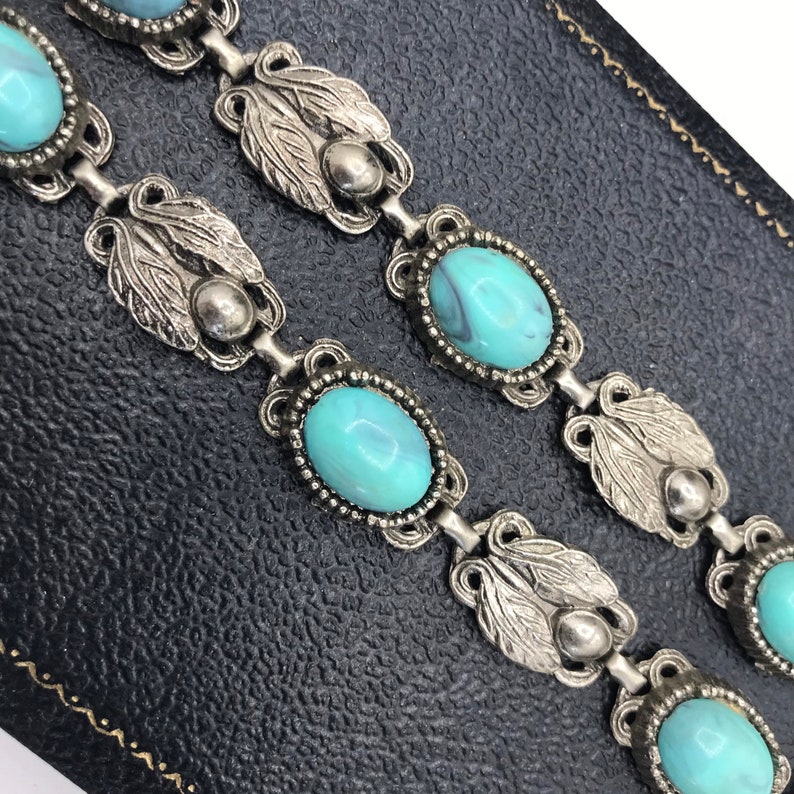 Op de afbeelding: Close-up van een vintage zilverkleurige armband met ovale turquoise stenen. De armband heeft sierlijke blad- en kraaldetails. Het ontwerp is klassiek en elegant. De armband wordt gepresenteerd op een zwarte ondergrond.