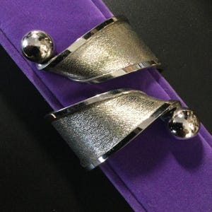May include: A pair of silver-toned cuff bracelets with a textured finish and a polished ball accent. The bracelets are displayed on a purple surface, showcasing their modern design and metallic sheen. The cuff bracelets are a stylish accessory.