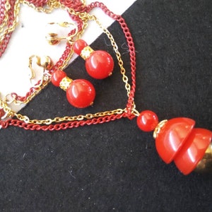 May include: A red and gold necklace and earrings set. The necklace has three chains, one red and two gold. The pendant is a red, layered, bell-shaped charm. The earrings are clip-on and feature two red beads.