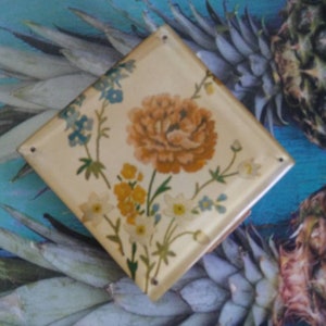 May include: A square, cream-colored tile featuring a central orange flower surrounded by smaller yellow and blue floral illustrations. The tile has a smooth, glossy finish and small holes at the corners, displaying a botanical design on its surface.